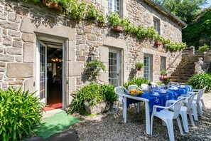 A stone building with a dining area set up outside, featuring a table with chairs and a blue tablecloth.