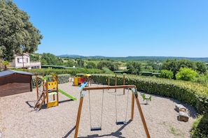 Ein Spielplatz mit Schaukeln und Klettergeräten, umgeben von einer Hecke und Blick auf ferne Hügel.