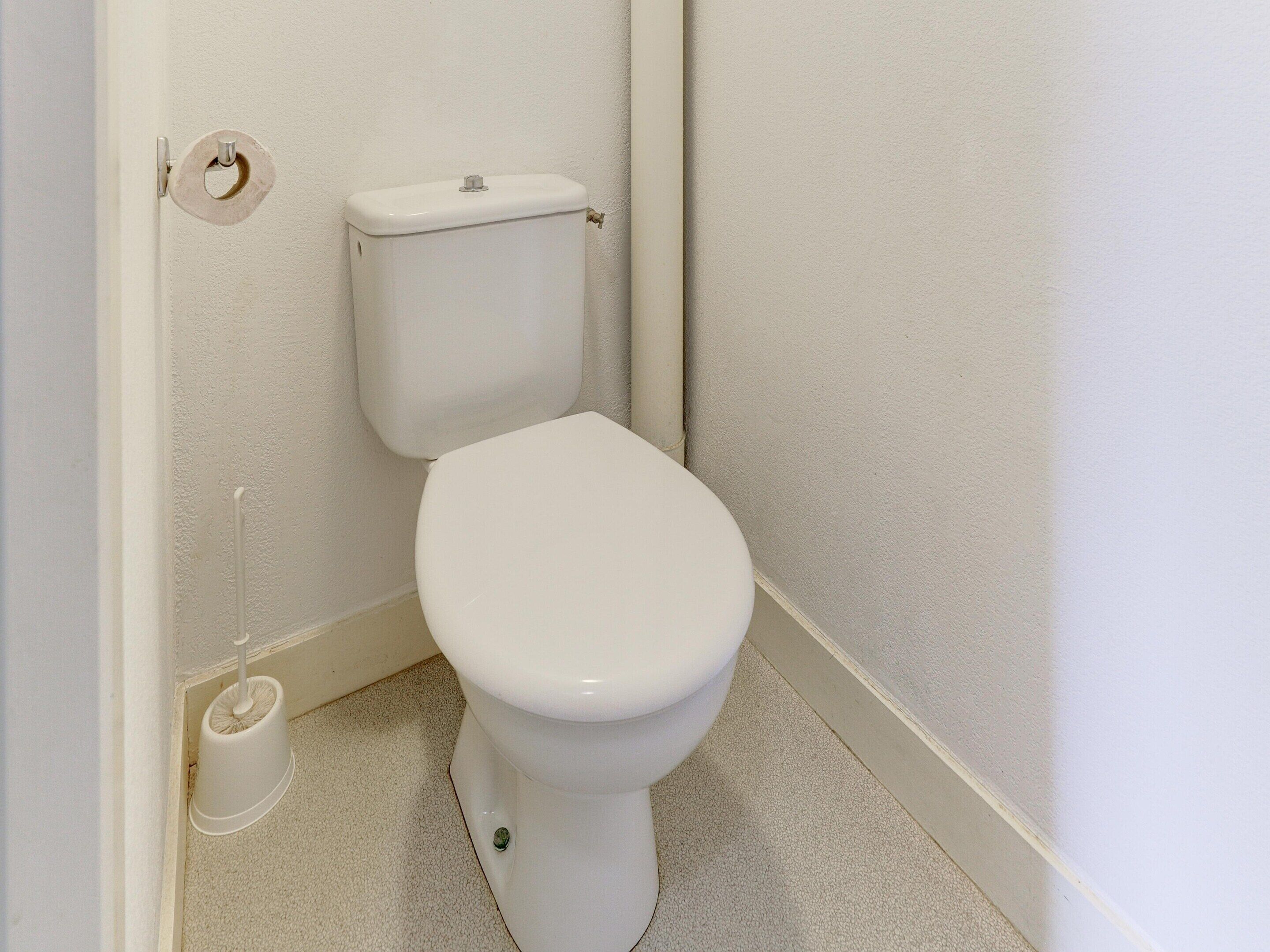 Un inodoro blanco en una esquina, con un dispensador de papel higiénico y una tubería visible en la pared.