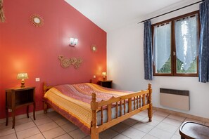 A bedroom with a wooden bed, two bedside tables with lamps, a window with curtains, and wall decorations. - Holiday Home “Le Cyprès - La Fontinelle” (Bessas)