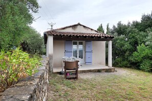 Ein kleines Haus mit Ziegeldach, einer Veranda mit Tisch und Stühlen sowie ein Garten mit Steinmauer und viel Grün.