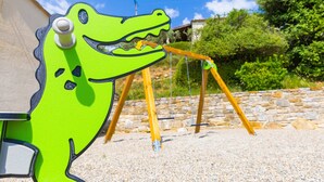 A green dinosaur-shaped swing set in a garden with stone walls and greenery. - Holiday Home “Le Cyprès - La Fontinelle” (Bessas)