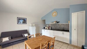 A compact living space with a kitchenette, a dining area with a wooden table and chairs, a gray sofa, and a framed picture on the wall. - Holiday Home “Le Cyprès - La Fontinelle” (Bessas)
