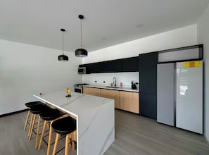 Uma cozinha moderna com ilha central, banquetas, luminárias pendentes e uma geladeira.