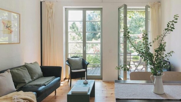 A living room with a sofa, a chair, a coffee table, and a dining table with a pitcher of flowers. - Art Deco villa with Havel views in historic Werder (Werder (Havel))