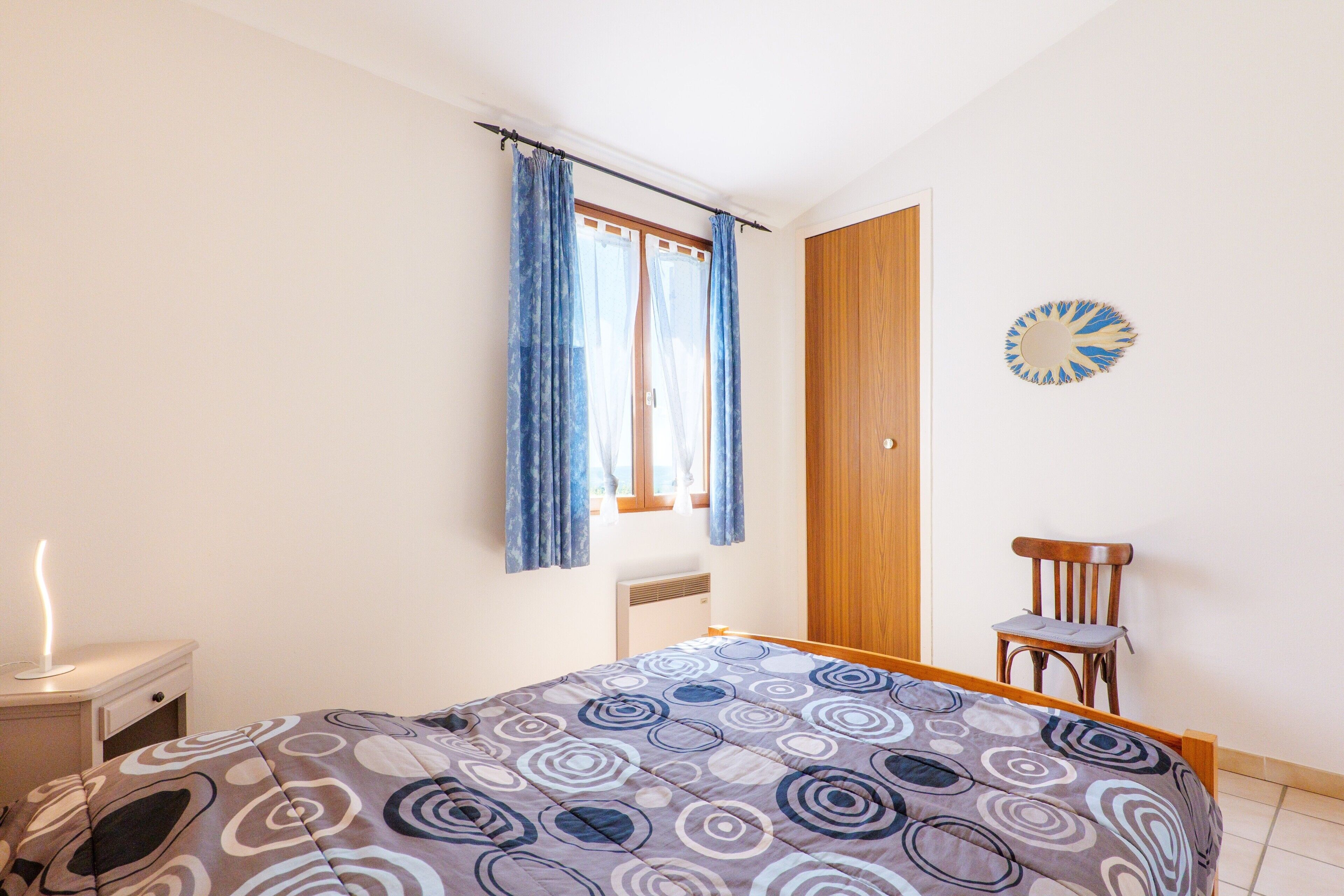 A bedroom with a bed, a chair, a nightstand, a window with curtains, and a wall decoration.