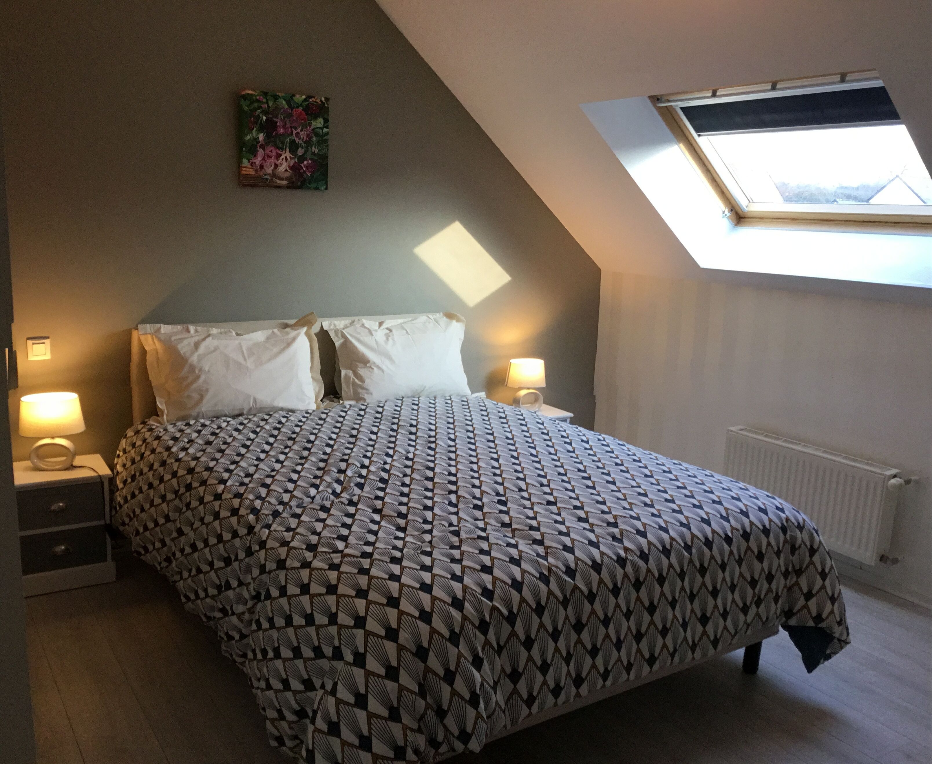 A bedroom with a sloped ceiling, a skylight, a patterned bedspread, two bedside lamps, a nightstand, and a radiator.
