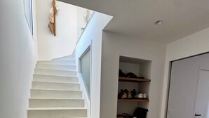 Un escalier aux marches blanches menant à un palier, avec une main courante en bois et une étagère à chaussures où sont rangées différentes paires de chaussures.