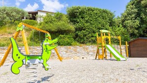 Un’area giochi con altalene e uno scivolo, circondata da un muro in pietra e vegetazione.