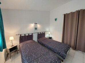 Ein Zimmer mit zwei Betten, einem Nachttisch mit Lampe, einer Wandregal und einem Fenster mit Vorhängen.