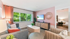 Uma sala de estar moderna com sofá, mesa de centro e TV, uma janela grande com vista para vegetação e uma parede de destaque em tom rosa.