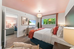 A bedroom with a bed, a TV, a desk, a chair, a window with a view, and a door leading to another room. - Boca Raton Apartment with King Bed Across the Beach. Near Mizner Park. (Boca Raton)
