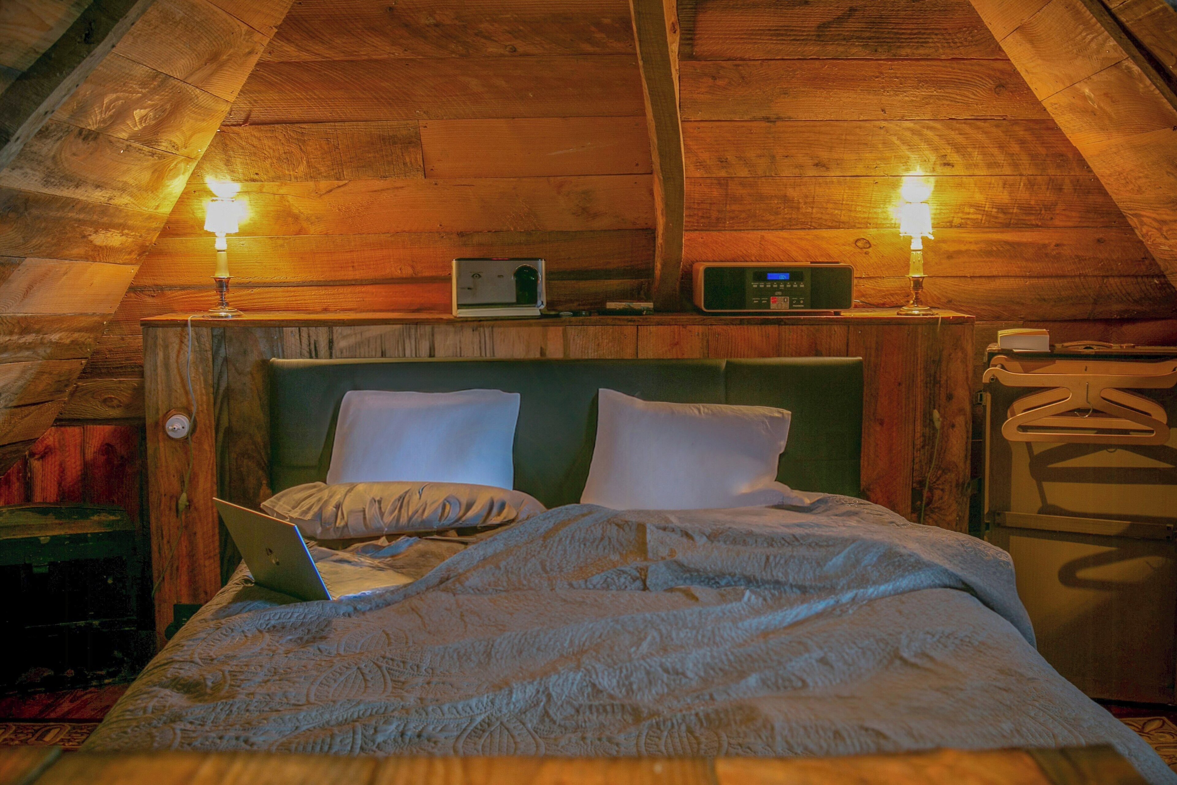 Une chambre mansardée chaleureuse, avec un plafond en pente en bois, un lit avec un ordinateur portable, deux lampes de chevet et une radio posée sur une étagère.