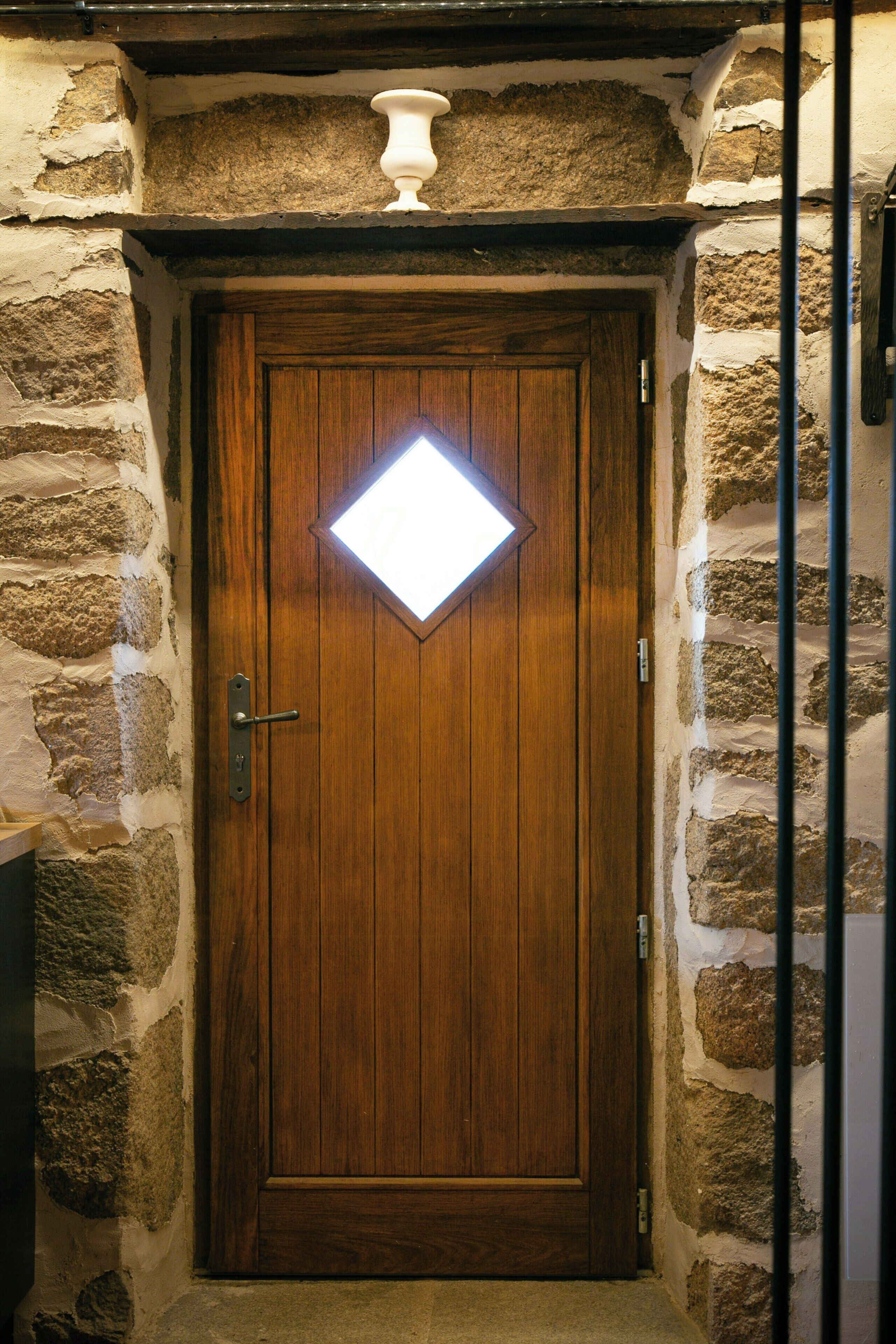 Une porte en bois ornée d’une fenêtre en forme de losange, encastrée dans un mur de pierre et surmontée d’un portail en métal.