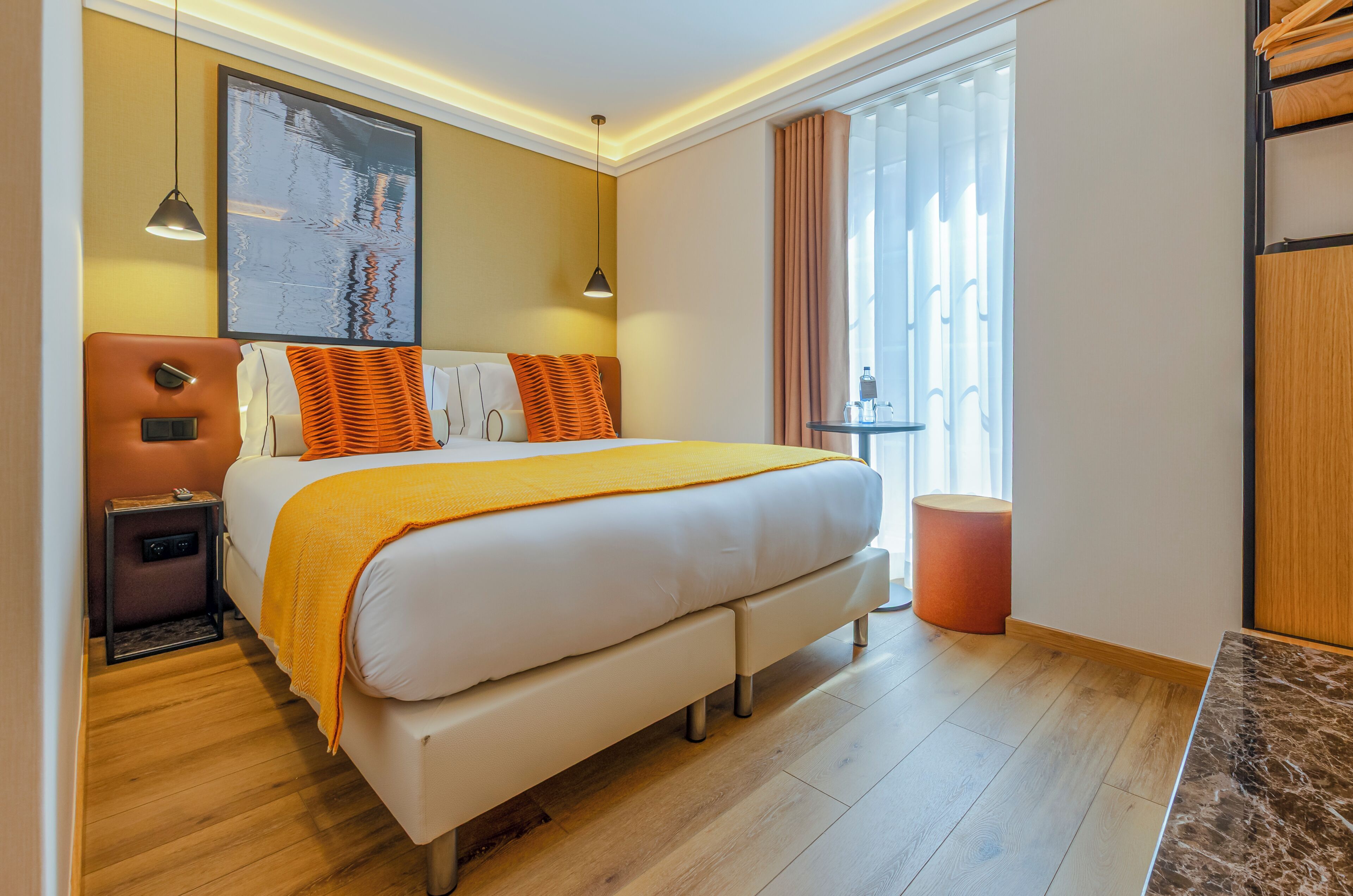Habitación de hotel con una cama grande, mesitas de noche, un tablero auxiliar, una obra de arte colgada en la pared, una ventana con cortinas y piso de madera.