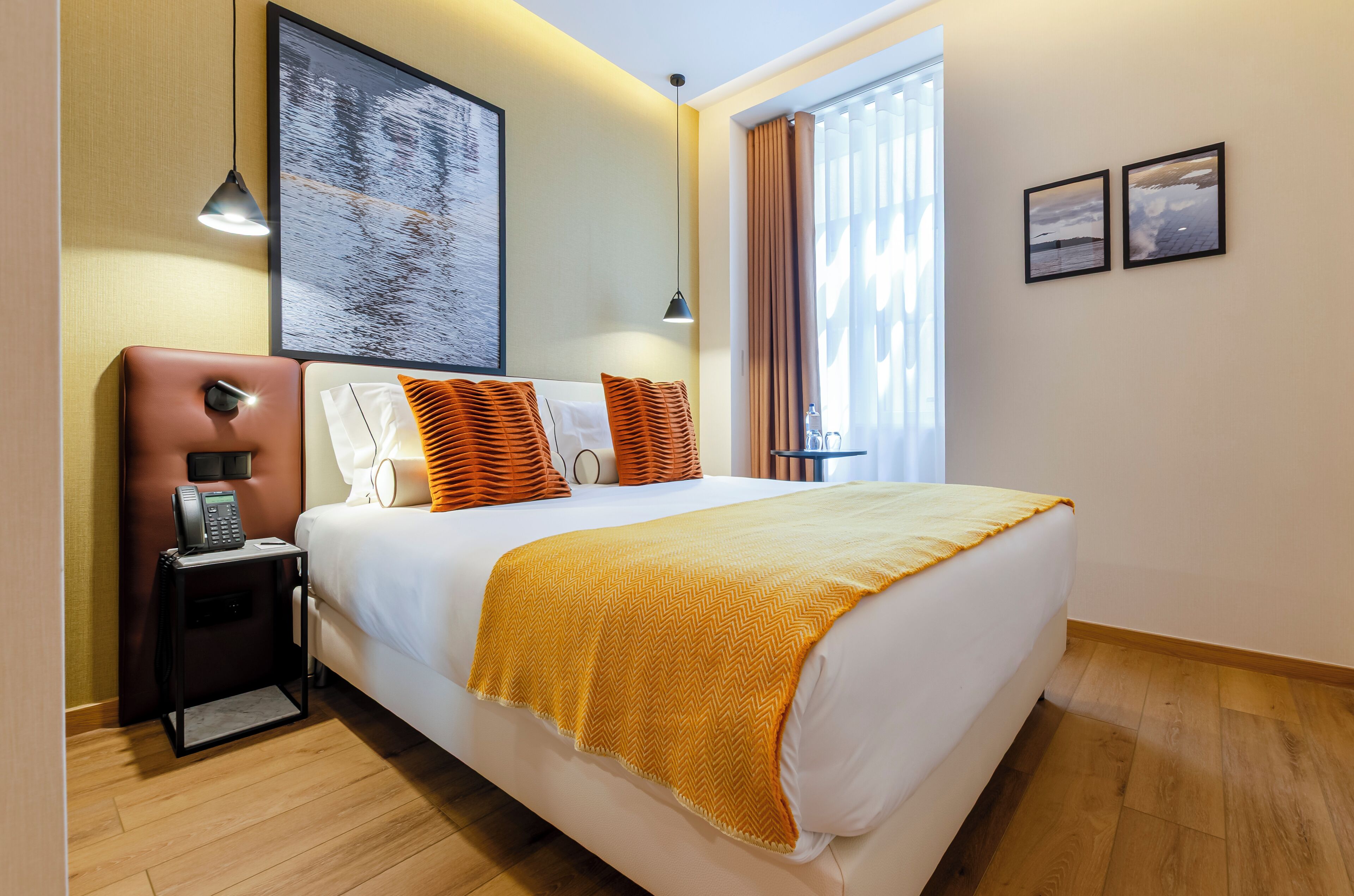 A hotel room with a bed, bedside table, lamp, artwork, and a window with curtains.