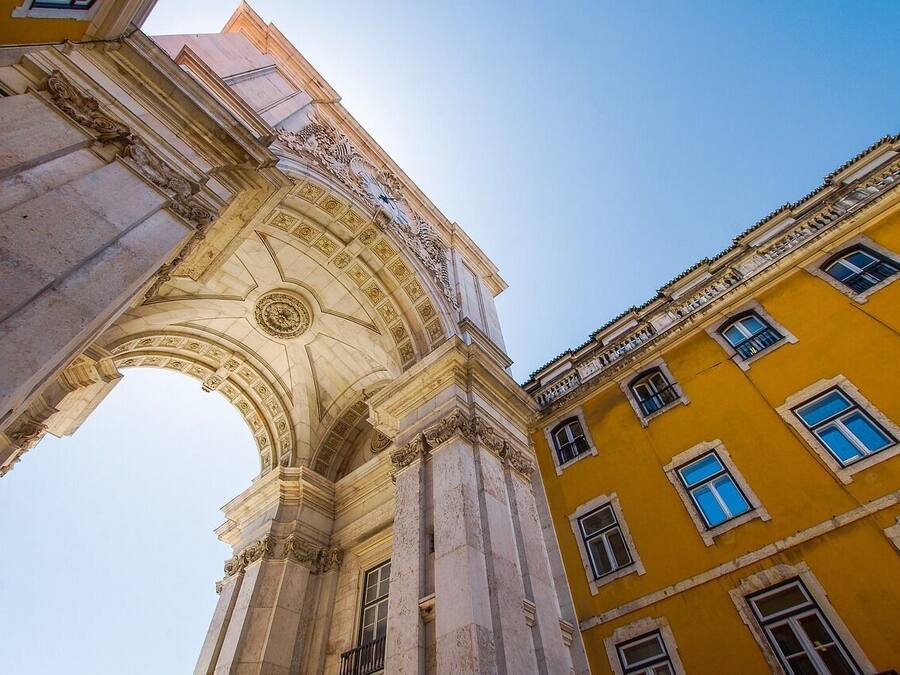 Un arco arquitectónico con elementos de diseño clásico, junto a un edificio amarillo con múltiples ventanas.