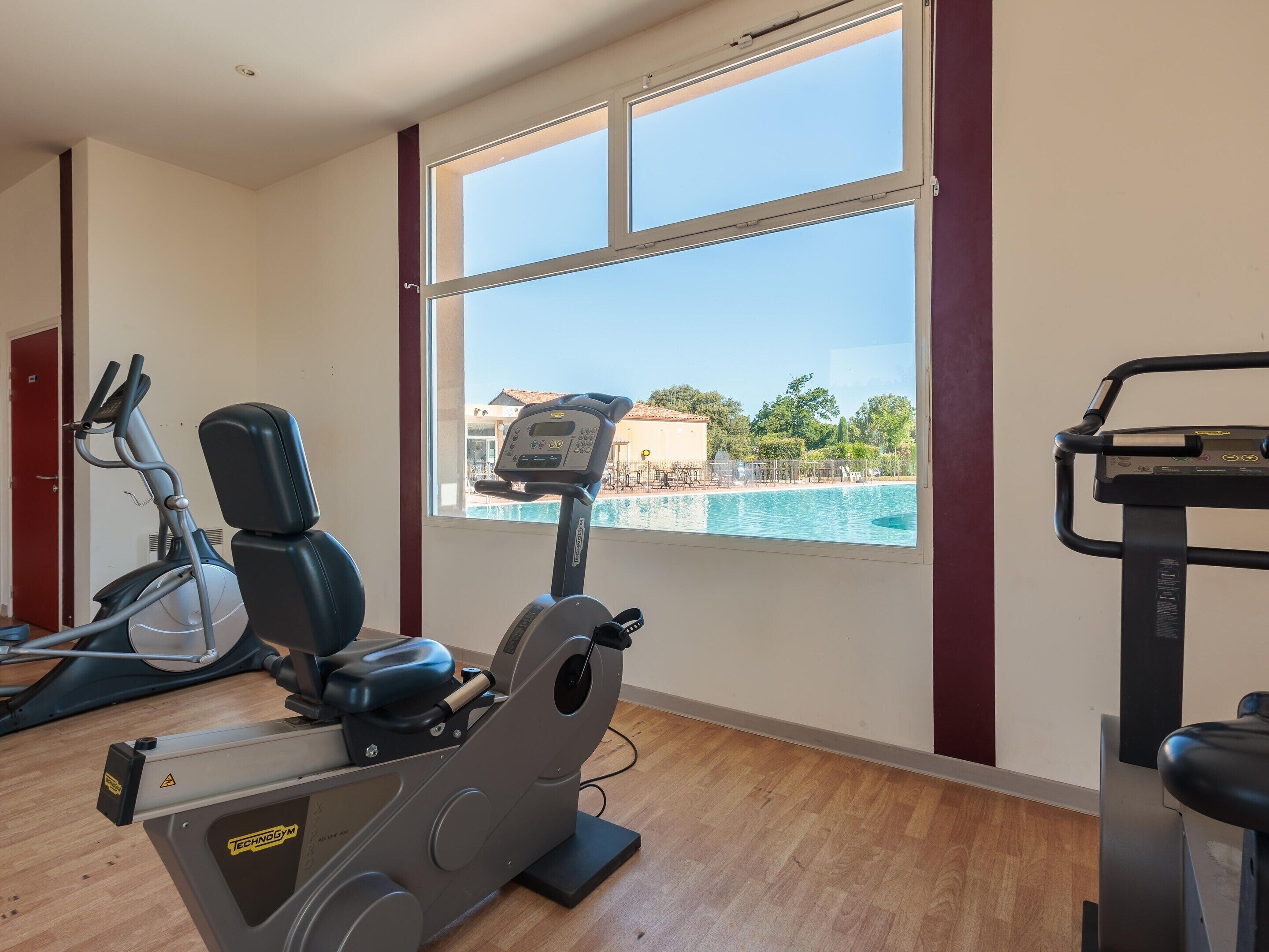 A gym with exercise equipment, a window with a view of a pool and buildings.