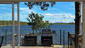 Katettu terassi, jossa on grilli ja tuoli, ja josta avautuu näkymä järvelle, jonka rannalla kasvaa puita ja jonka yllä on kirkas taivas.