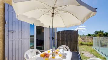 Mesa al aire libre con una sombrilla blanca, sillas blancas y una mesa de desayuno con frutas, jugo y una botella.