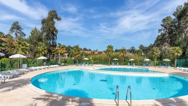 Une piscine rectangulaire, bordée d’une terrasse carrelée, entourée de chaises longues et de parasols, avec des arbres et un ciel dégagé en arrière-plan.