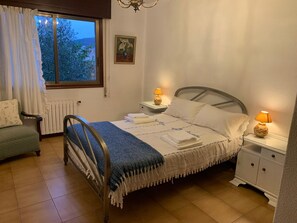 A bedroom with a bed, bedside tables, a chair, a radiator, a window with curtains, and a painting on the wall.
