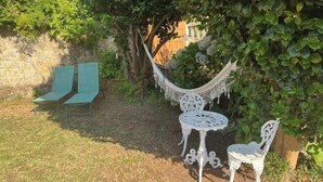 Eine Hängematte, zwei weiße Metallstühle und ein kleiner runder Tisch in einem Garten mit Grünpflanzen und einer Steinmauer.