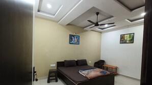 A bedroom with a bed, a nightstand, a chair, a ceiling fan, and artwork on the walls. - Hotel Shriji Nikunj (Mathura)