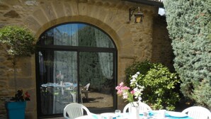 A stone building with a large arched doorway, outdoor seating with white plastic chairs and a table, and a potted plant. - Holiday Home 'La Bergerie' with Shared Pool, Private Terrace and Wi-Fi (Fontarèches)