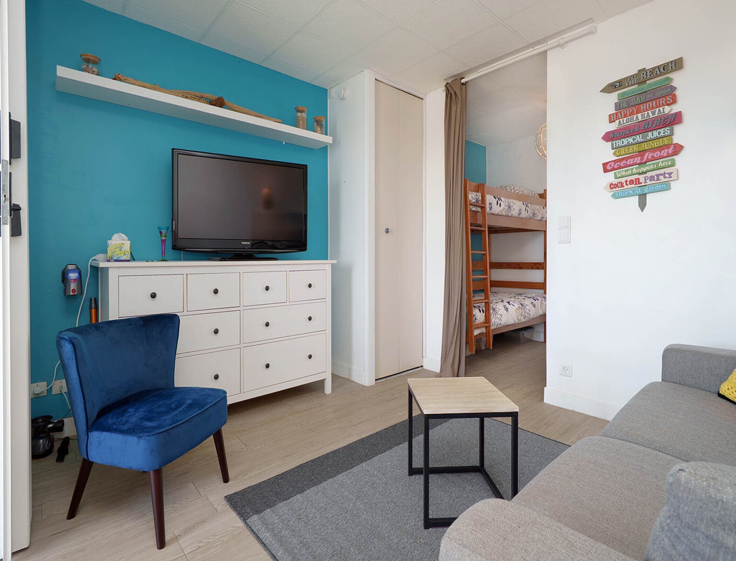 A room with a TV on a white cabinet, a blue chair, a grey sofa, a bunk bed, and a wall with a sign.