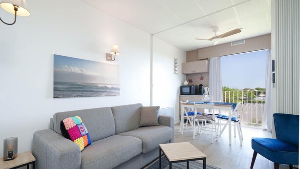 Een moderne woonkamer met een grijze bank, een klein houten bijzettafeltje, een tv op een meubel en een schilderij van de oceaan, plus een balkon met uitzicht.