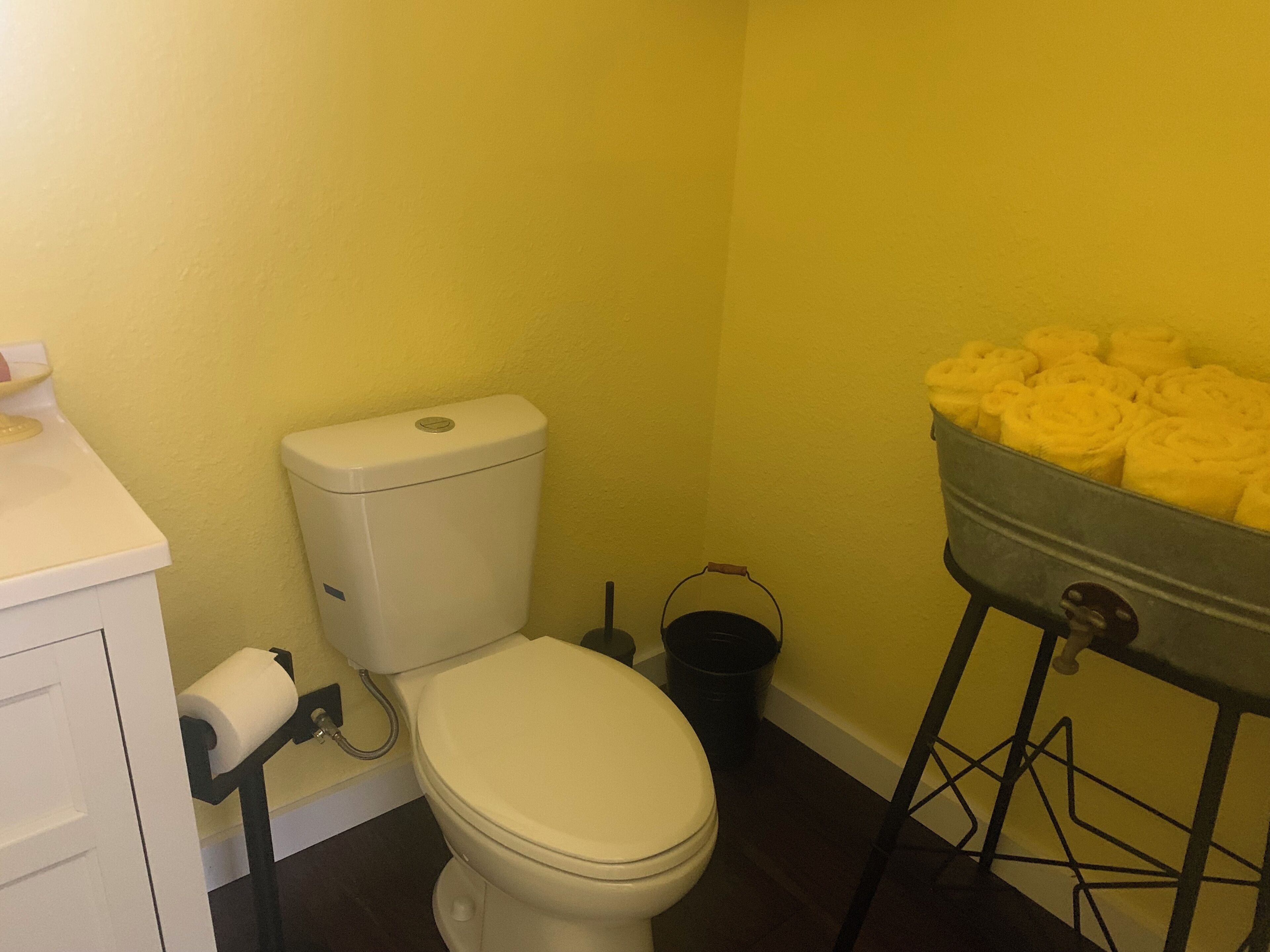 Een badkamer met een toilet, toiletpapier, een zwarte emmer en een metalen bak met handdoeken.