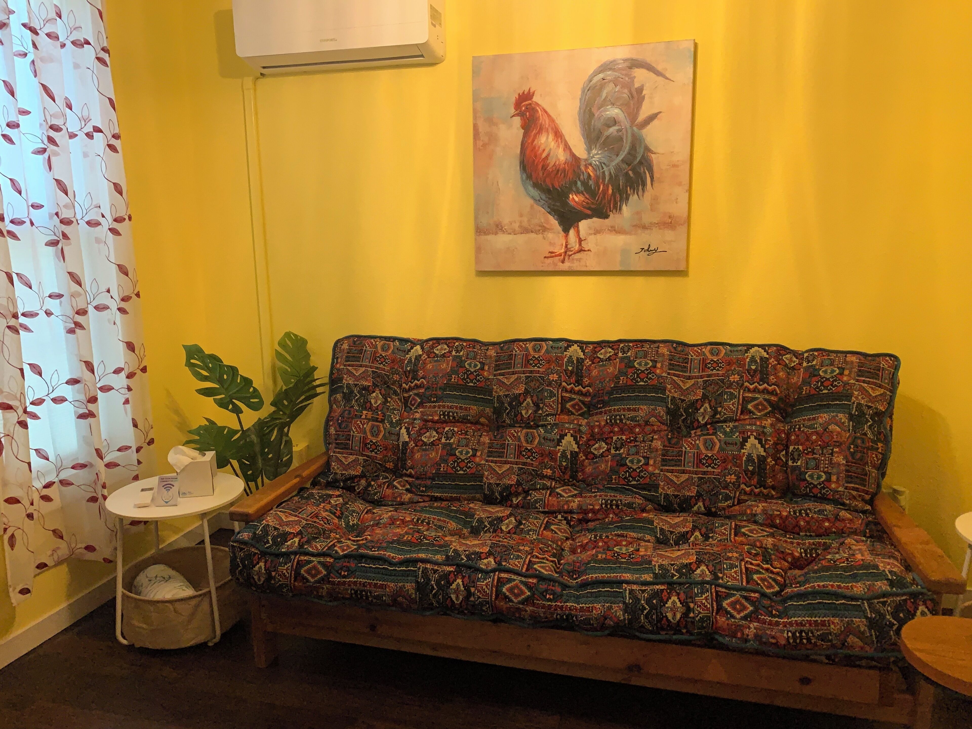Une pièce avec un canapé à motifs, un tableau représentant un coq, une plante en pot et une petite table sur laquelle se trouve un panier.