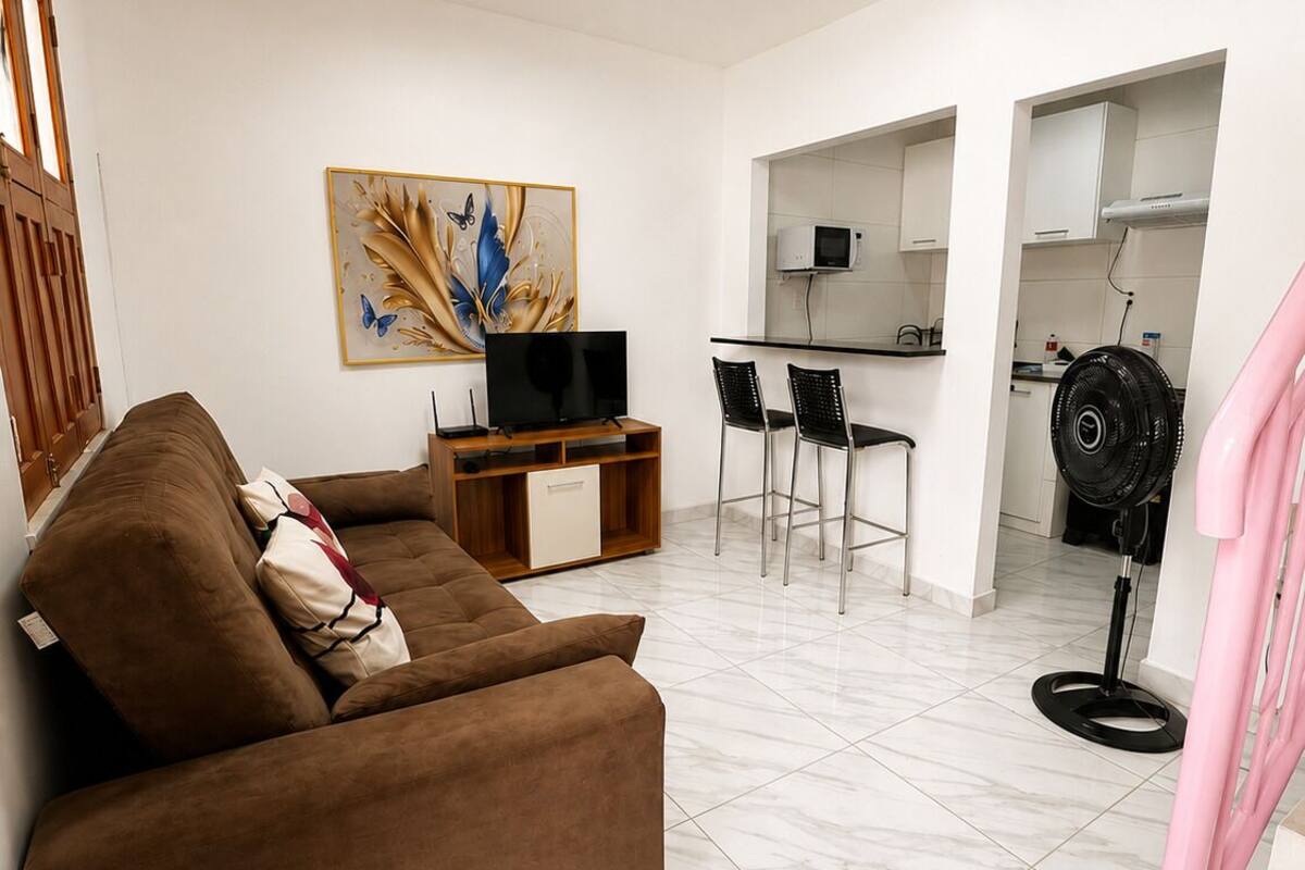 A living room with a sofa, TV, and artwork; a kitchen with a dining area; and a staircase.
