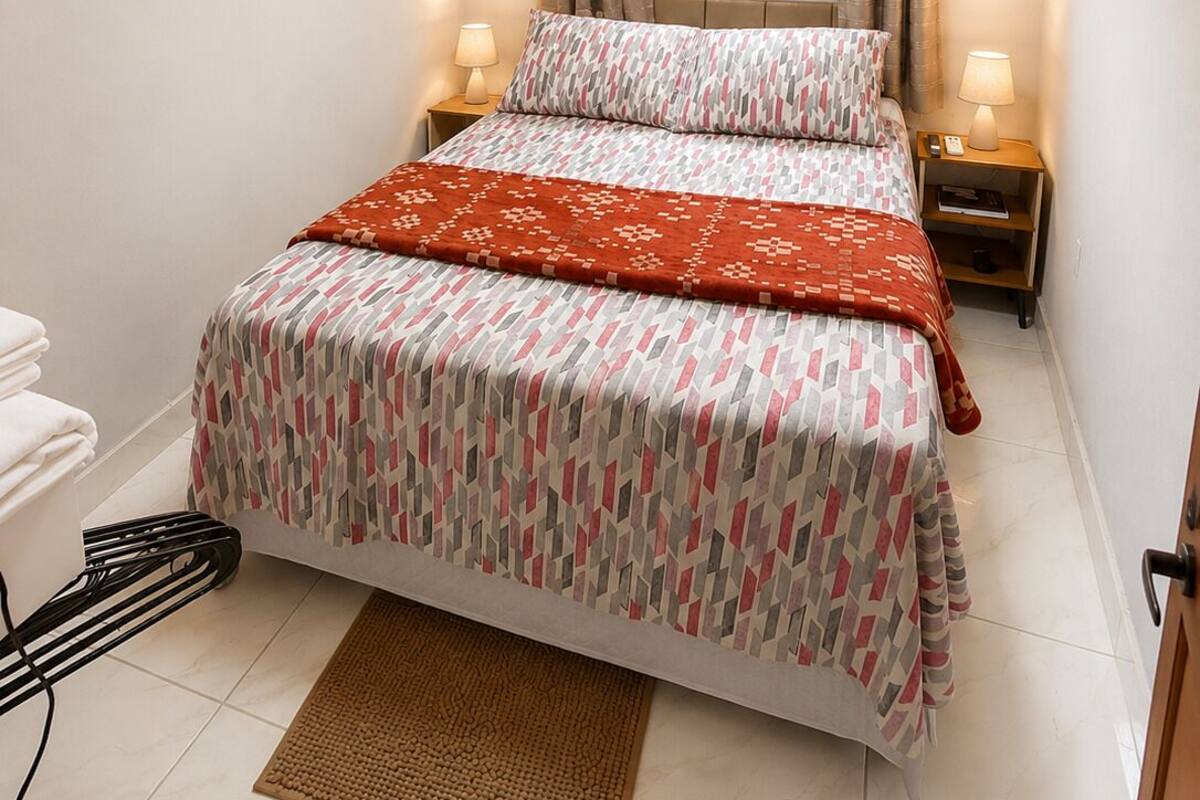 A bedroom with a bed, two bedside lamps, a window with curtains, an air conditioning unit, and a bedside table.