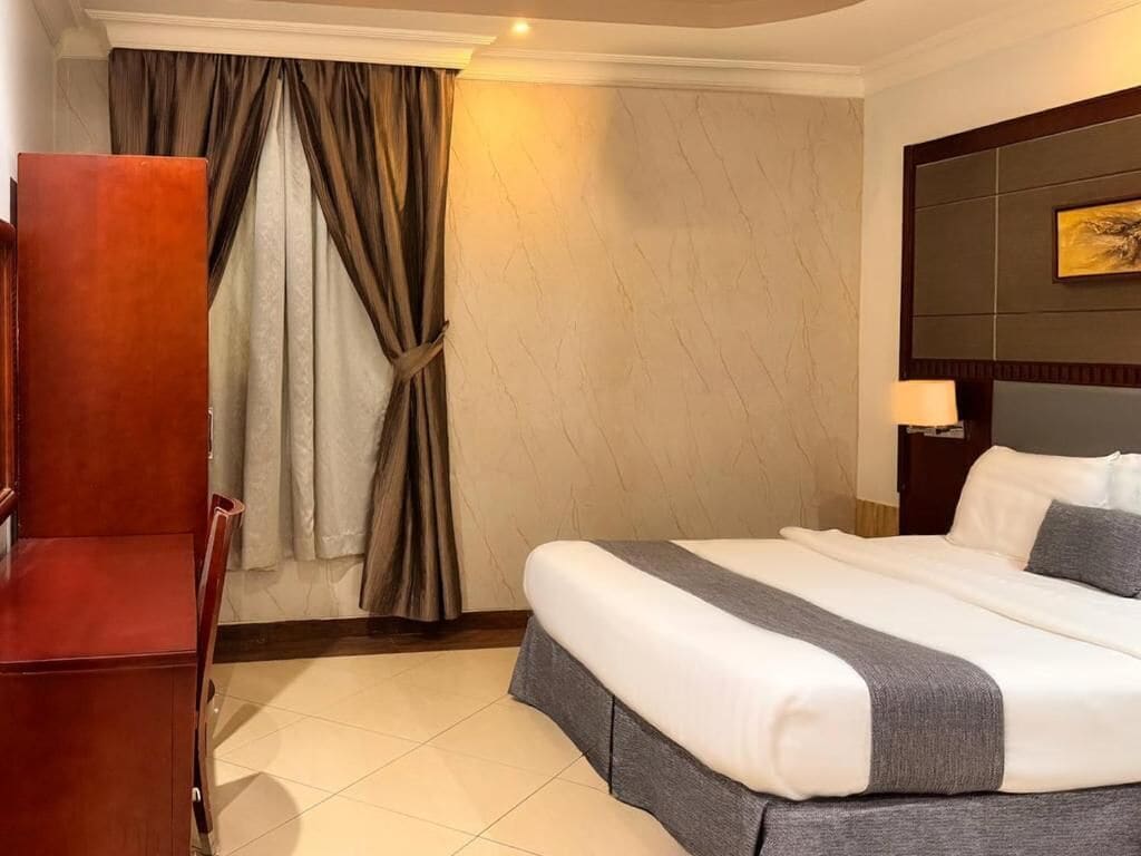 Una camera d'albergo con un letto, una sedia di legno, un comodino, una lampada e una tenda.