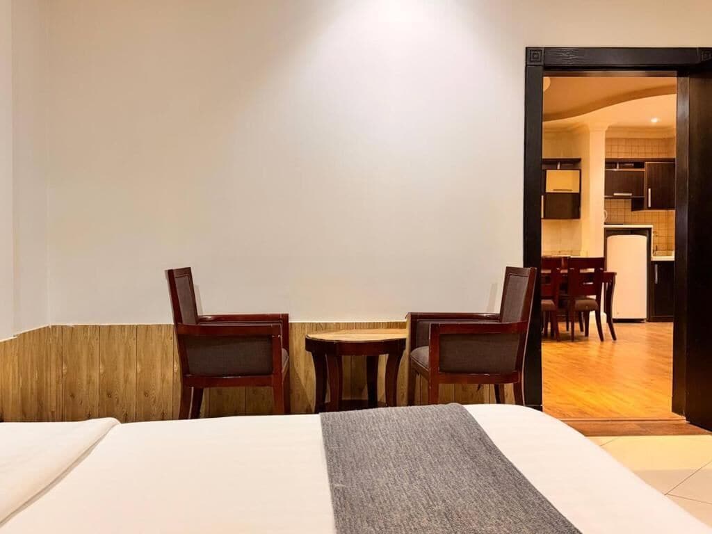A hotel room with a bed, two chairs, a small table, and an open doorway to a kitchen area.