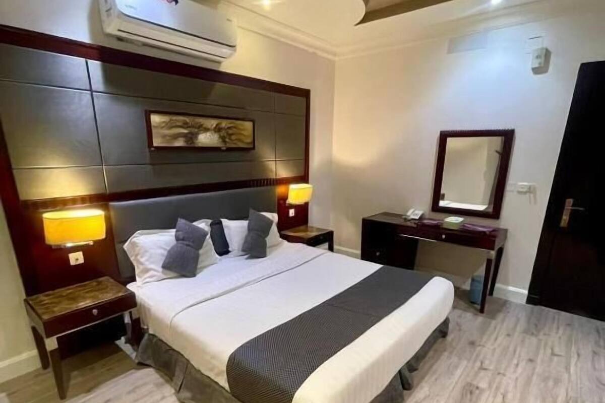 A hotel room with a bed, bedside lamps, a nightstand, a dresser, a mirror, and an air conditioning unit.