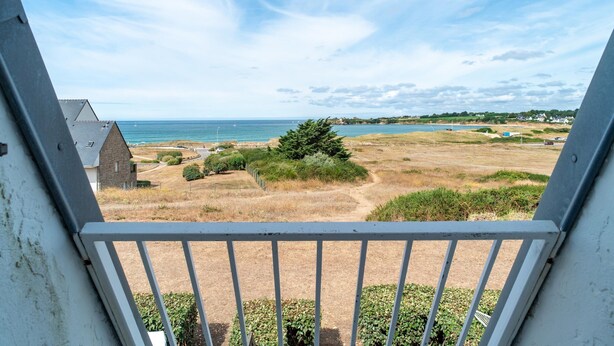 A balcony with a view of a coastal landscape, including a grassy field, a few trees, and a distant body of water. - Guidel Sea View Apartment for 5 (Guidel)
