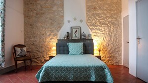 A bedroom with a stone wall, a bed with a patterned bedspread, a chair with a cushion, and a nightstand with a lamp.