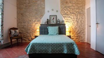 Een slaapkamer met een stenen muur, een bed met een beddeken met een patroon, een stoel met een kussen en een nachtkastje met een lamp.