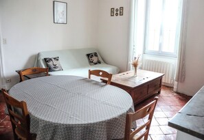 A room with a round table, wooden chairs, a sofa with patterned cushions, a radiator, and a window with curtains.