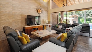 A modern living room with a flat-screen TV, leather sofas, a coffee table, and large windows offering a view of a house and greenery. - Casa Avándaro (Valle de Bravo)