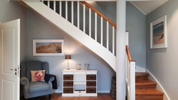 Una escalera con peldaños de madera y barandillas blancas, un sofá gris con un cojín rojo, una consola blanca con cajones y un cuadro en la pared.