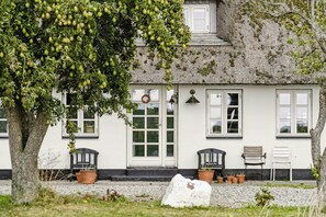 En hvid bygning med stråtækt tag, et stort træ med grønne frugter, og to krukker med planter på verandaen.