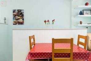 Ein Essbereich mit einem Tisch, der mit einer roten Tischdecke mit weißen Punkten bedeckt ist, zwei Holzstühlen, einer Vase mit roten Tulpen und einem an der Wand hängenden Bild von Keramik.