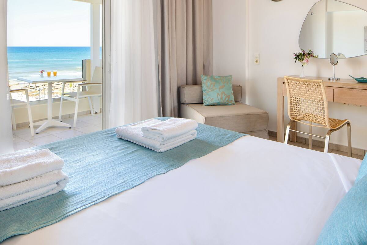 A hotel room with a bed, a desk, a chair, a view of the beach, and a window with curtains.