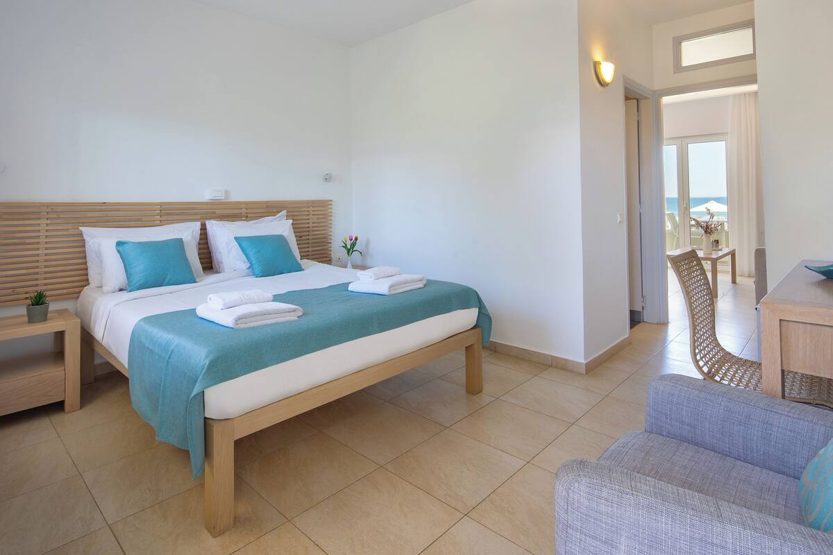 A hotel room with a bed, a chair, a small table, and a view of the sea through a window.