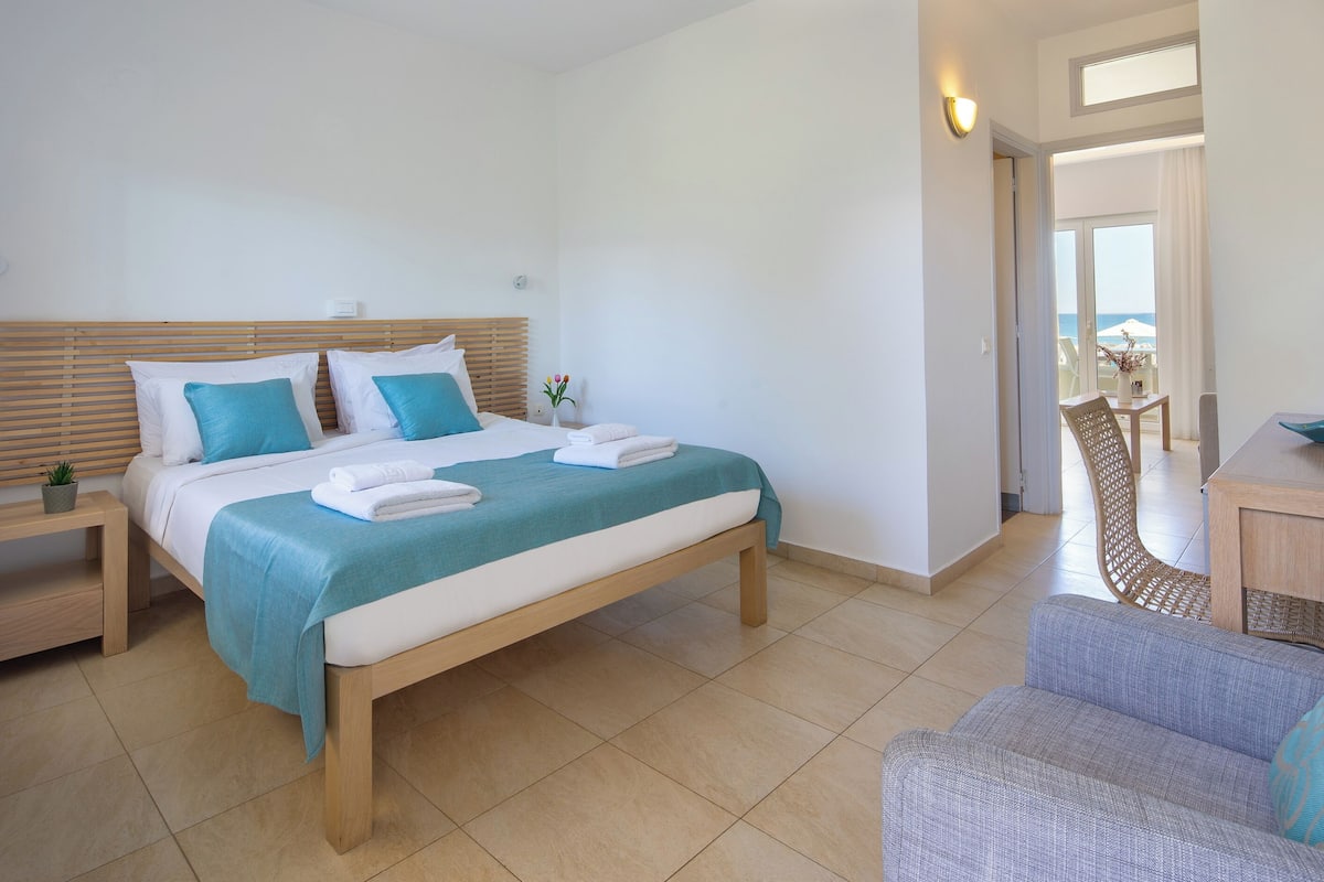 A hotel room with a bed, a chair, a small table, and a view of the sea through a window.