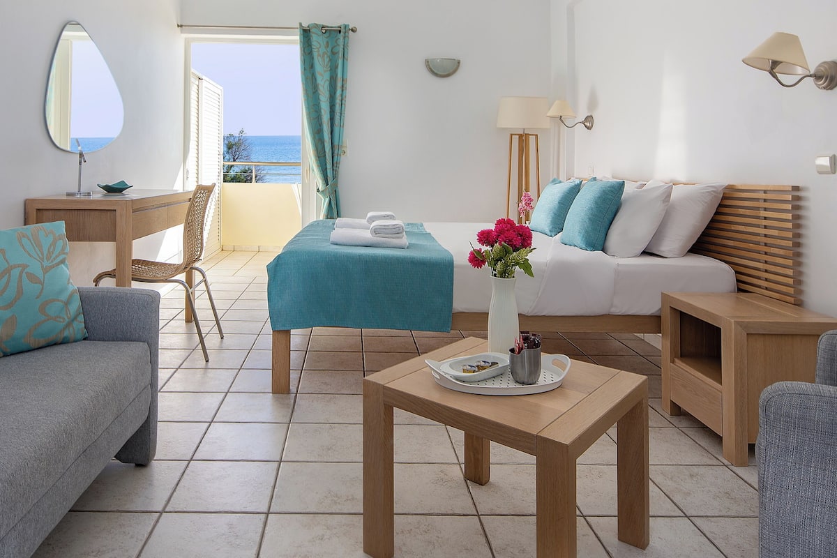 A hotel room with a bed, a desk, a chair, a small table, and a view of the sea.